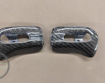 May include: Two carbon fiber automotive trim pieces. Each piece has a curved shape with an oval opening and a metal bracket. The carbon fiber has a black and gray woven pattern. The background is a light tan color.