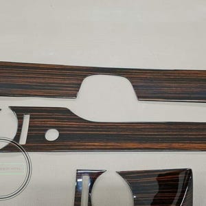 Puede incluir: Un conjunto de molduras interiores para automóviles con un patrón de vetas de madera. Las piezas son de varias formas y tamaños, con un acabado brillante. La veta de la madera presenta tonos marrón oscuro y rojizo. Las molduras son probablemente para el salpicadero o los paneles de las puertas de un coche.