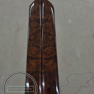 May include: Two matching, glossy, wood-grain panels with a rich, dark brown finish. The panels have a vertical, elongated shape with rounded tops. The wood grain features a swirling pattern. The panels are likely automotive interior trim.