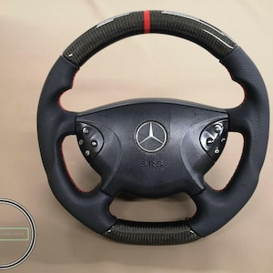 Op de afbeelding: Zwart en koolstofvezel stuurwiel met rode stiksels en een rode streep aan de bovenkant. Het midden heeft het Mercedes-Benz logo en de letters "SRS".