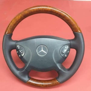 May include: A black and wood-effect steering wheel featuring the Mercedes-Benz emblem. The wheel has integrated controls and the letters "SRS" are visible below the emblem. The rim is wood-effect.