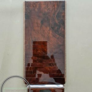 May include: A rectangular, wood-grain panel with a glossy finish. The panel has a dark brown, brick-like pattern and a chrome-colored, horizontal bar at the bottom. The text "wood and carbon work" is in a circular logo in the lower left corner.