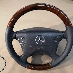 Könnte beinhalten: Ein schwarzes Lenkrad mit Holzmaserung und dem Mercedes-Benz-Logo in der Mitte. Das Lenkrad hat Bedienelemente auf beiden Seiten und einen strukturierten Griff. Die Holzmaserungsbereiche sind dunkelbraun.