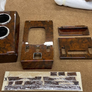 May include: A set of car interior trim pieces with a glossy, dark wood grain finish. Includes a cup holder, center console panel, and other components. The pieces are arranged on a brown surface.
