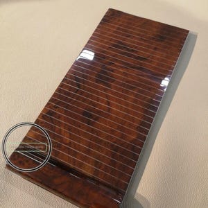 Puede incluir: Panel rectangular brillante con veta de madera marrón oscuro. El panel presenta una serie de líneas paralelas finas y claras en su superficie. La veta de la madera tiene un patrón natural y arremolinado. Probablemente una pieza de moldura interior automotriz.