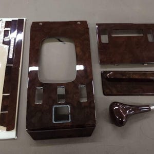 May include: A collection of car interior trim pieces in a dark wood grain finish. The set includes a center console panel, door trim, and a gear shift knob. The pieces are arranged on a neutral surface.