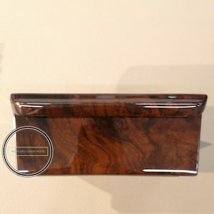 Puede incluir: Un objeto rectangular de madera brillante con un rico patrón de vetas marrón oscuro. El objeto tiene un acabado pulido y una ligera curva en el borde superior. El texto "wood and carson works" está en un logotipo circular en el lateral.