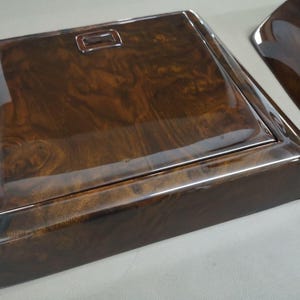 May include: Two pieces of dark brown, glossy wood trim with a burl grain pattern. One piece is rectangular with a recessed rectangular detail. The other piece is a curved shape. The wood has a high-gloss finish.