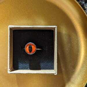 May include: A silver ring with an orange and black eye-shaped stone, presented in a small cardboard box with a black interior. The box sits on a gold-colored, round tray.