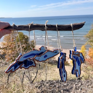 May include: Five blue and white layered lake outline ornaments hanging from a driftwood branch. The ornaments are suspended by twine and are in the shape of the Great Lakes. The background is a blurry view of a lake and shoreline.