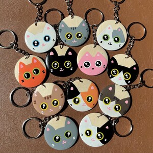 Puede incluir: Una colección de once llaveros circulares de madera, cada uno con una cara de gato de dibujos animados con ojos grandes. Los gatos están representados en varios colores, incluyendo naranja, negro, blanco, gris, marrón y tricolor.