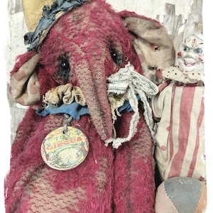 Pode incluir: Um brinquedo de elefante de pelúcia vermelho vintage com um chapéu azul e amarelo, um colar listrado azul e branco e uma etiqueta redonda amarela que diz "Miniature Toy Circus". O elefante está de pé ao lado de uma boneca de palhaço vintage com um traje listrado vermelho e branco e uma bola.