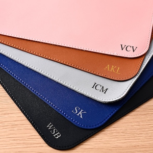 May include: A stack of personalized mouse pads in various colors: pink, brown, gray, blue, and black. Each pad has a different set of initials in the corner. The pads are made of a smooth material and are stacked on a wooden surface.