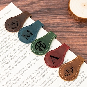 May include: A collection of leather magnetic bookmarks in various colors, including brown, teal, green, burgundy, and tan. Each bookmark features a unique initial or monogram design, with a stitched border. The bookmarks are placed on the pages of a book.