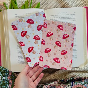 May include: Two decorative cards with a mushroom pattern. The cards feature red mushrooms with white polka dots and tan stems, set against a white and pink background with gold stars. The cards are held open over an open book.