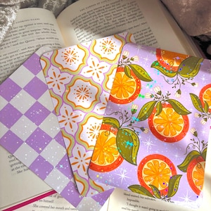 May include: Three decorative bookmarks with distinct designs are shown on an open book. One has a purple and white chequered pattern, another features an orange and green floral design, and the third showcases orange slices. The bookmarks are on a soft, textured surface.