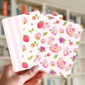May include: Three square cards with various patterns are held in front of a blurred bookshelf. The cards feature pink stripes, strawberries, teapots, and floral designs in pink, red, and green. The cards are held by a person with pink nail polish.