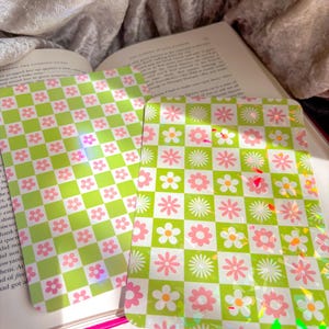 May include: Two decorative cards featuring a green and white chequered pattern, embellished with pink and white floral designs. The rectangular cards are made of a shiny material. The overall theme is floral and decorative.