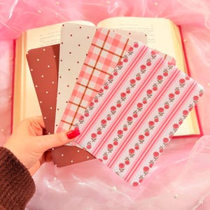 May include: A set of four rectangular bookmarks featuring distinct patterns: one dark red with white polka dots, one cream with brown polka dots, one with a pink and white gingham check, and one with vertical pink stripes and small red rose motifs.
