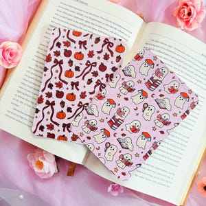 May include: Two rectangular bookmarks resting on an open book. One bookmark features a pattern of orange pumpkins, red apples, acorns, and maroon ribbons. The other bookmark displays a pattern of white ghosts reading books and small stars.