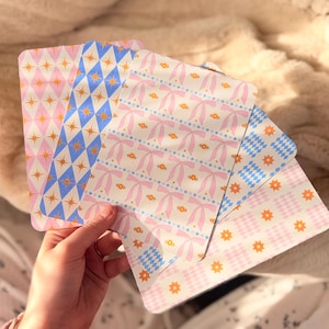 May include: A hand holds four decorative cards with different patterns. The cards display repeating designs of bows, stars, and diamonds in pastel pink, blue, and gold. The cards are rectangular with rounded corners.