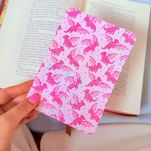 May include: A rectangular bookmark featuring a repeating pattern of bright pink dragons with spread wings on a light lavender background. The surface is covered in a holographic star and sparkle effect that reflects light across the design.