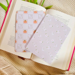 May include: Two decorative rectangular bookmarks are placed on an open book. One bookmark has a repeating pattern of orange pumpkins and white flowers on a grey checkered background. The other has a pattern of white flowers on a grey background.