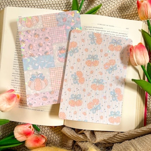 May include: Two decorative cards with pumpkin designs. One card features a patchwork pattern with pumpkins, while the other has a repeating pumpkin and bow pattern. The cards are set on an open book with tulips.