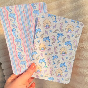 May include: Two notebooks with a pastel ocean theme. One has a striped pattern with dolphins and seahorses. The other has a repeating pattern of dolphins, seahorses, shells, and stars. Both have a glittery finish.
