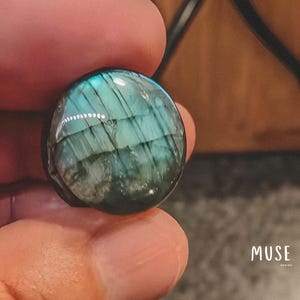 Puede incluir: Una gema de labradorita pulida y redonda, con tonos iridiscentes azules y verdes. La piedra se sostiene entre los dedos, mostrando sus patrones naturales y cualidades reflectantes. La palabra "MUSE" es visible en la esquina inferior derecha.