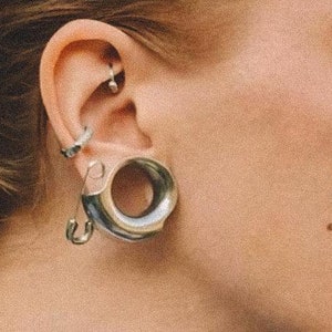 Puede incluir: Una persona con un aro de plata en la oreja y un anillo grande de plata en el lóbulo de la oreja. El anillo es un círculo grande con un círculo más pequeño en el centro. La persona tiene una coleta corta y castaña. El texto "MUSE DESIGN" es visible en la imagen.