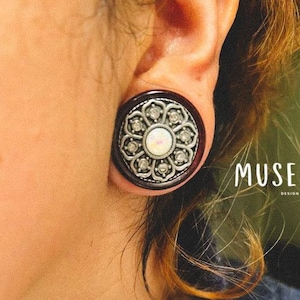 Pode incluir: Um close-up de uma orelha com uma dilatação de orelha de madeira escura e prata. A dilatação tem um design decorativo com uma opala branca no centro. O texto "MUSE DESIGN" é visível no canto inferior direito da imagem.