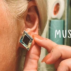 May include: A diamond-shaped earring with a gold frame and an iridescent abalone shell inlay is being held up to an ear. The earring has a black center and is labeled "MUSE DESIGN".