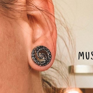 May include: A black, circular earring with a spiral design, resembling a tentacle, is worn in an ear. The earring is detailed with small, circular accents around the edge. The word "MUSE" and the word "DESIGN" are printed in black.