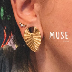 May include: Gold leaf-shaped earrings with a textured surface. The earrings are worn in the earlobe and helix piercing. The earrings are from the brand MUSE Design.