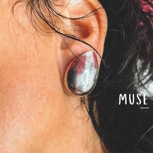 以下が含まれることがあります： 耳に大きな楕円形の銀色のイヤリングをしている人のアップ。イヤリングは滑らかで反射しています。