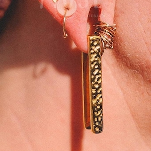 Könnte beinhalten: Ein Ohr mit mehreren goldenen Ohrringen und Ohrsteckern. Die Ohrringe umfassen einen Ring, eine gebogene Stange und einen langen, strukturierten Stab-Ohrring.