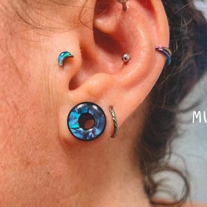 May include: Close-up of an ear with multiple piercings. The ear features a large, round, iridescent blue and black plug, a small green hoop, a silver stud, a crescent-shaped stud, and a curved, multi-colored hoop. The word "MUSE" is visible.