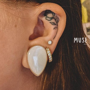 Peut inclure: Une paire de boucles d'oreilles en forme de larme blanche avec des montures dorées. Les boucles d'oreilles sont portées dans les lobes inférieurs de l'oreille. L'oreille droite a également un petit clou de diamant et une boucle d'oreille dorée avec de petits diamants.