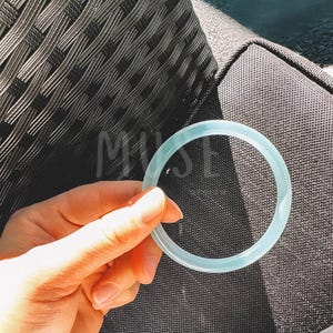 K&ouml;nnte beinhalten: Ein hellblauer Silikonring, in einer Hand gehalten, vor einem dunkelgrauen, strukturierten Hintergrund.