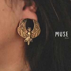 May include: A gold-toned earring featuring a detailed design of a moth with a skull head. The earring is in the shape of a crescent moon.