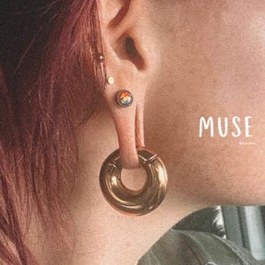 May include: A person with pink hair wearing a gold hoop earring with a rainbow coloured stone in their ear. The text "MUSE DESIGN" is visible in the image.