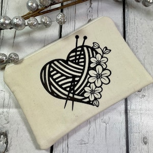 May include: A white canvas zippered pouch with a black and white graphic of a heart-shaped ball of yarn with knitting needles and flowers.