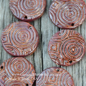 May include: Close-up of several round, reddish-brown ceramic beads with a glossy finish. Each bead features a concentric circle and square pattern design. The beads have small holes for stringing. The text "www.CapturedMoments.Etsy.com" and "Made in Texas, USA" are visible.