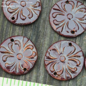 May include: Four round ceramic tiles with a brown glaze and a white floral design. The tiles have two holes each and are made in Texas, USA.