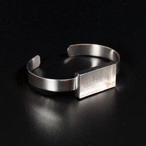 May include: A silver bracelet with a rectangular, open-faced bezel. The bezel is smooth and has a slightly textured surface.