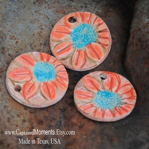 May include: Three ceramic ornaments with a floral design. Each ornament features a stylized flower with a turquoise center and orange petals. The ornaments have a hole at the top for hanging.