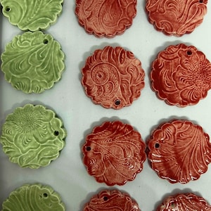 May include: Overhead shot of ceramic ornaments in shades of green and red. Each ornament has a scalloped edge and a detailed floral or swirl design. A small hole is at the top of each ornament.