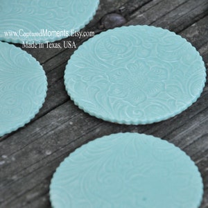 May include: Several round, light blue coasters with a floral embossed design. The coasters have a scalloped edge and are arranged on a wooden surface. The text "www.CapturedMoments.Etsy.com Made in Texas, USA" is visible.