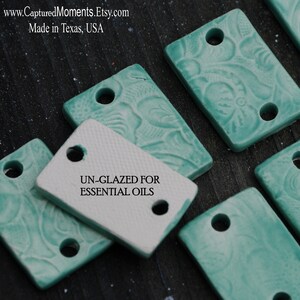 May include: A close-up of several small, rectangular ceramic tiles with a teal glaze and a floral pattern. The tiles have two holes in them and are labeled "UN-GLAZED FOR ESSENTIAL OILS".